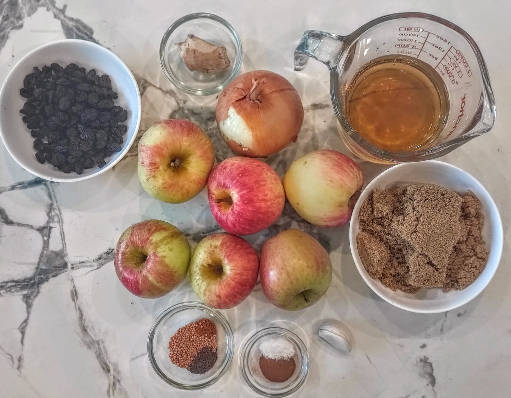An assortment of ingredients laid out on a marble countertop, including several apples, a bowl of black raisins, a piece of ginger, a measuring cup with liquid, an onion, and bowls of brown sugar and various spices.