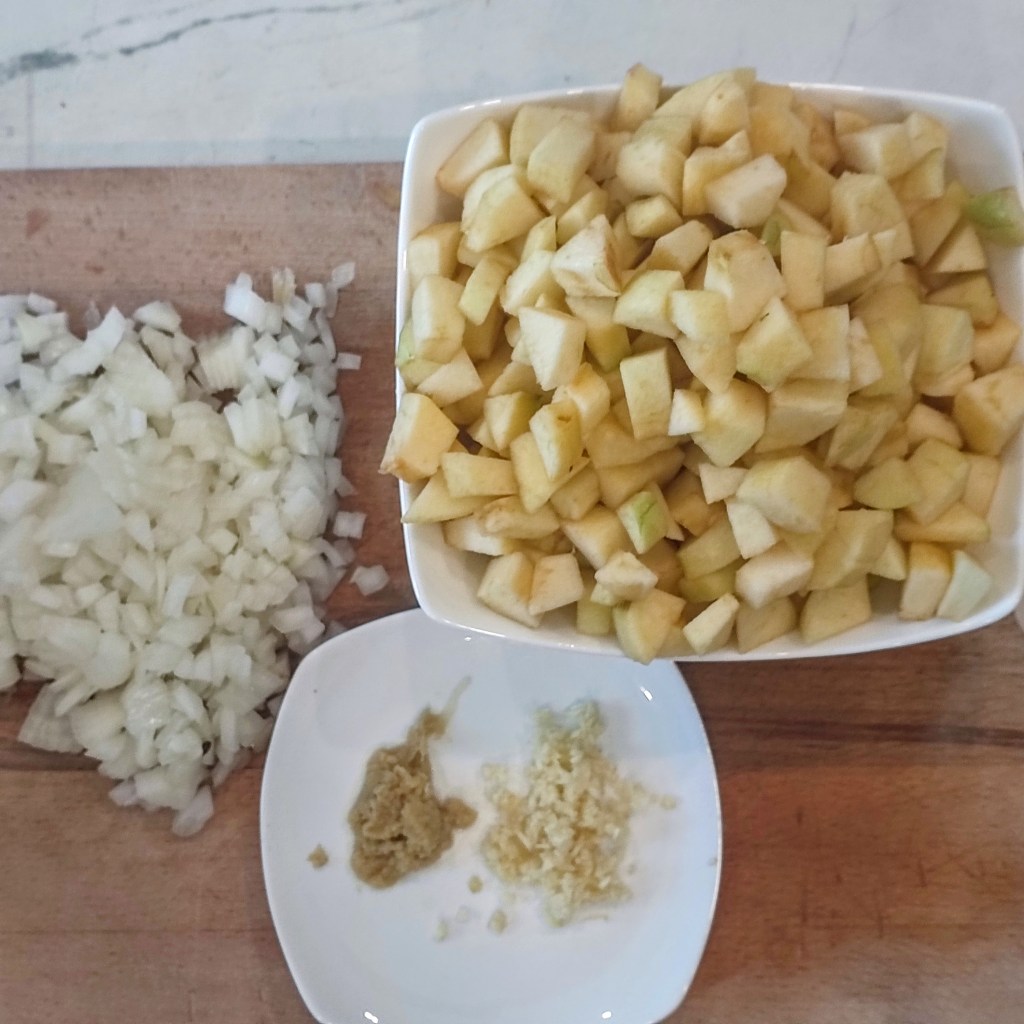 Chopped onions, diced apples in a bowl, and minced ginger and garlic on a plate, arranged on a wooden cutting board.