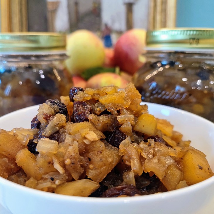 A bowl of fruit preserve made with diced apples, raisins, and spices, with jars of preserves and fresh apples in the background.